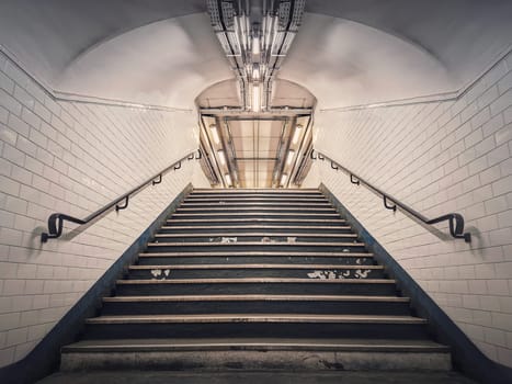 Subway corridor with white tile on walls and glowing lights above a the stairs passage. Symmetrical underground staircase