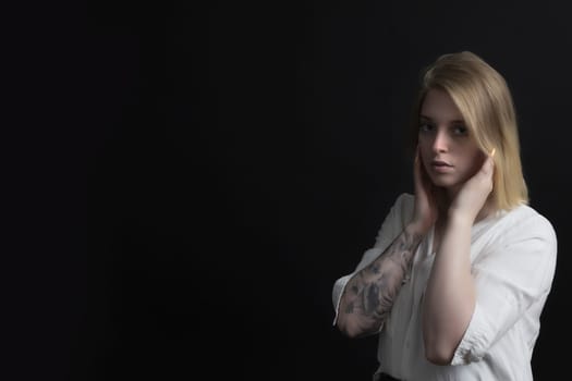 Low key portrait of blonde sad young woman weared in white shirt with tattoo on her hand.