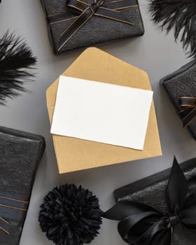 Blank card and envelope between Wrapped Gift Boxes and black feathers, mockup, copy space. Dark composition with horizontal paper card for Birthday, Anniversary or black party present