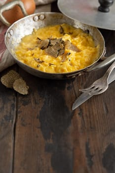 Scrambled eggs with sliced fresh black truffles from Italy served in a a frying pan near fork and knife on wood, close up, copy space. Italian cousine, gourmet breakfast