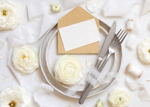 Wedding table setting with card and envelope near cream roses and white silk ribbons top view, mockup. Romantic scene with horizontal table place card and pastel flowers. Valentines, Spring or Mothers day concept