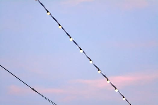 A row of glowing string lights suspended on a cable, set against a gradient sky transitioning from light blue to pinkish hues. The scene captures the serene and magical atmosphere of twilight.