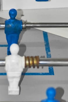 Close-up of foosball player figures, one white and one blue, mounted on metal rods with gold springs. The scene captures the dynamic and fun nature of table soccer games.
