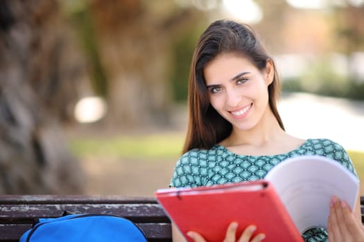 Happy student holding notes looks at camera sitting on a bench in a park