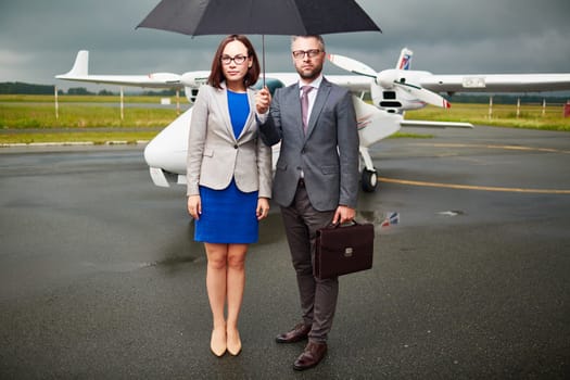 Young businessman holding umbrella over himself and his partner on background of airplane