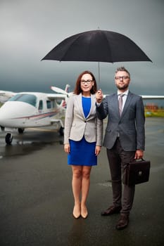 Two confident colleagues under umbrella looking at camera on overcast day