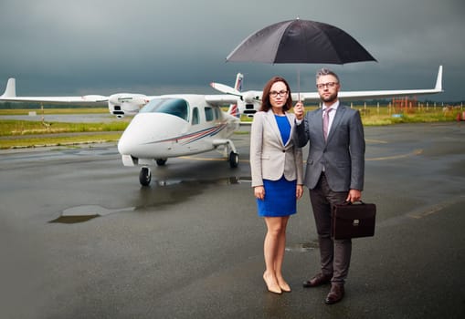 Two confident colleagues standing on asphalt on overcast day under umbrella