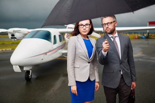 Elegant employees under umbrella looking at camera on rainy day on background of airplane