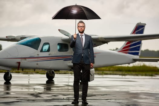 Serious businessman standing on asphalt on overcast day under umbrella