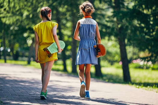 Backs of little girls with skateboards walking down park