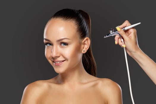 visagist making makeup for model with aerograph or airbrush on black background