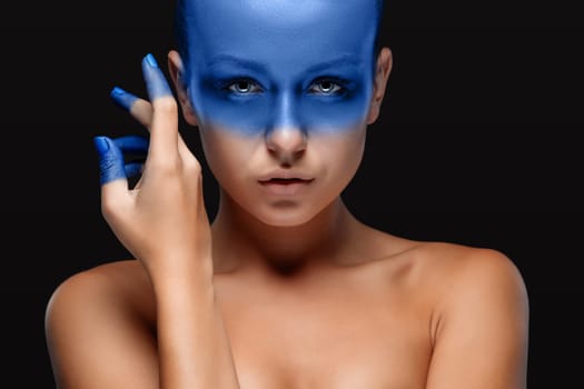 Portrait of a young woman who is posing covered with blue paint in the studio on a black background