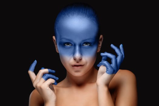 Portrait of a young woman who is posing covered with blue paint in the studio on a black background. girl&#39 s hands around the face and painted blue