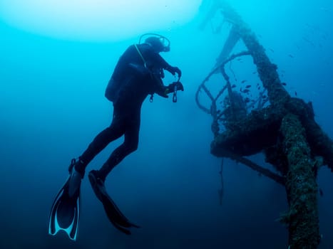 man in a black wet suit is diving a wreck into the ocean, the water is blue and the man is surrounded by fish