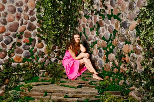 An adult woman sits on a stone plate near the wall in her cute summer dress, with wavy hair and a charming smile. She looks relaxed and content, enjoying the weekend outdoors.