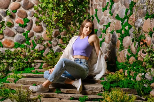 Cute young woman sits on stone path surrounded with old overgrown stone wall in the park