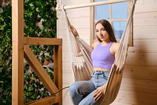 Cheerful woman is sitting in a hammock on the terrace of her country wooden house