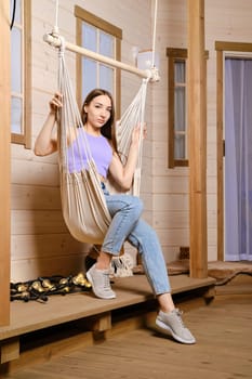 Cheerful woman is sitting in a hammock on the terrace of her country wooden house