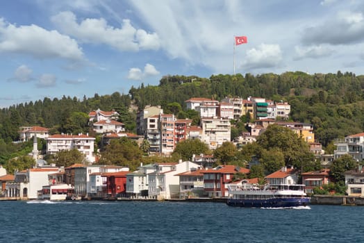 View from Bosphorus strait of the green mountains of the Asian side, with traditional houses, dense trees and a huge Turkish flag in the far end, in a summer day, Istanbul, Turkey