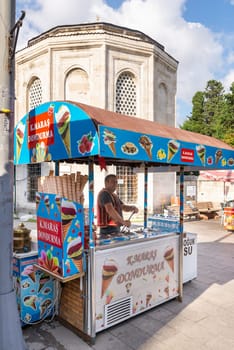 Istanbul, Turkey - September 1, 2022: Middle aged man selling ice cream, or dondurma on traditional Turkish fast food cart with historical tomb in the background at Eyupsultan district