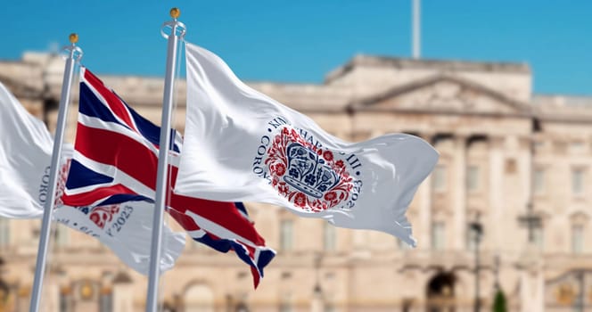 Flags with the emblem of the coronation of King Charles III and of UK waving. The emblem depicts flora of the four nations in the shape of St Edward’s Crown. 3d illustration render. Fluttering textile