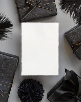 Blank card between Wrapped Gift Boxes, paper flowers and feathers, mockup, copy space. Dark composition with vertical paper card for Birthday, Anniversary or black party present