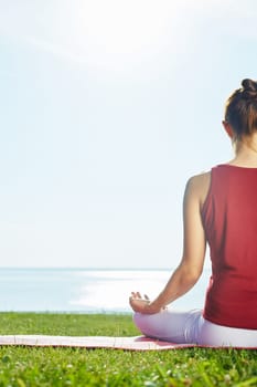 Relaxing woman sitting in pose of lotus on green grass by the seaside