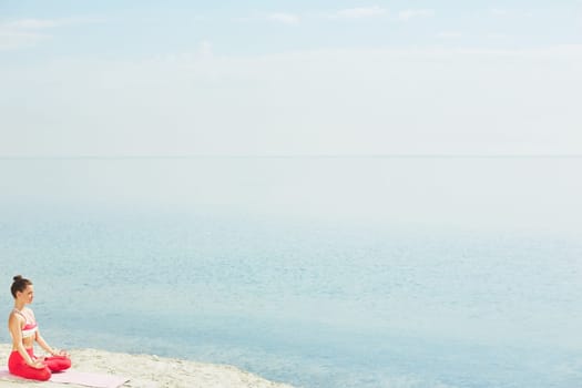 Sporty young woman sitting in pose of lotus on sand by water