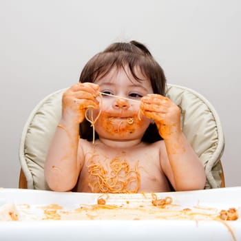 Happy baby having fun eating messy covered in Spaghetti holding Angel Hair Pasta red marinara tomato sauce.