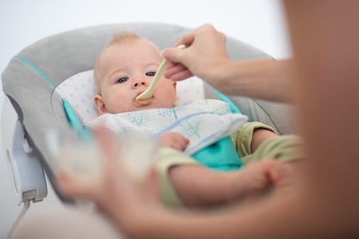 Mother spoon feeding her baby boy child in baby chair with fruit puree. Baby solid food introduction concept