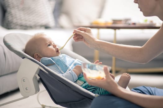 Mother spoon feeding her baby boy child in baby chair with fruit puree. Baby solid food introduction concept