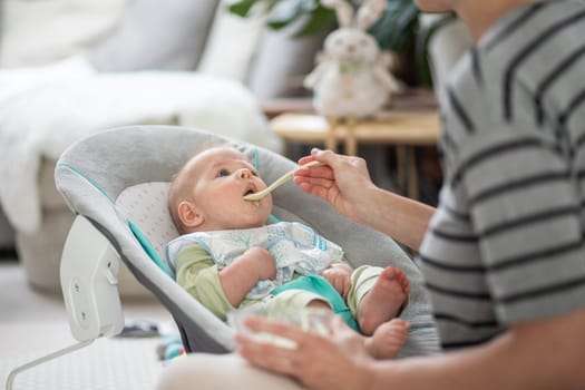 Mother spoon feeding her baby boy child in baby chair with fruit puree. Baby solid food introduction concept