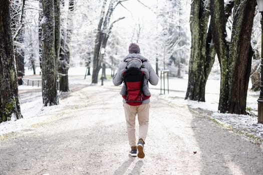 Rear view of sporty father carrying his infant son wearing winter jumpsuit and cap in backpack carrier walking in city park in winter.