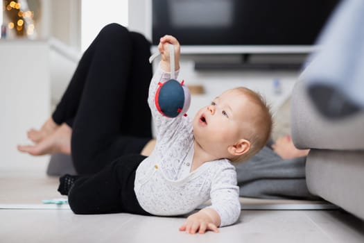 Happy family moments. Mother lying comfortably on children's mat watching and suppervising her baby boy playinghis in living room. Positive human emotions, feelings, joy.