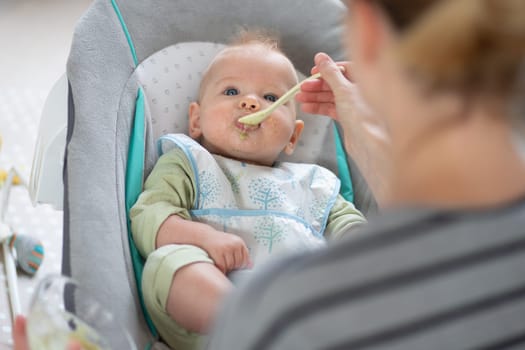 Mother spoon feeding her baby boy child in baby chair with fruit puree. Baby solid food introduction concept