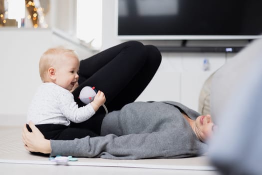Happy family moments. Mother lying comfortably on children's mat playing with her baby boy watching and suppervising his first steps. Positive human emotions, feelings, joy