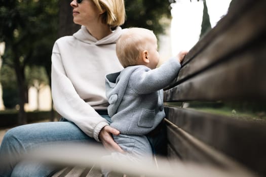 Young mother with her cute infant baby boy child sitting on bench in city park