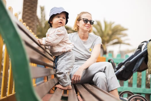 Young mother with her cute infant baby boy child on bench on urban children's playground on warm summer day