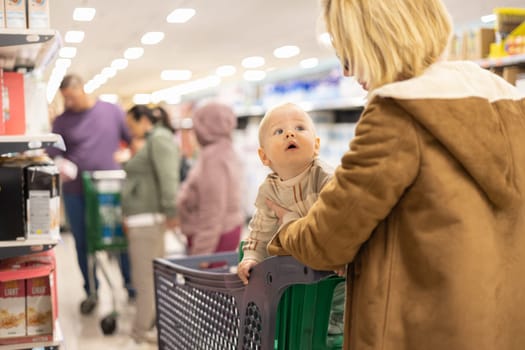 Casualy dressed mother choosing products in department of supermarket grocery store with her infant baby boy child in shopping cart