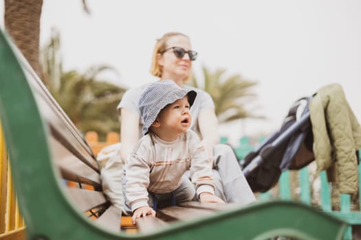 Young mother with her cute infant baby boy child on bench on urban children's playground on warm summer day
