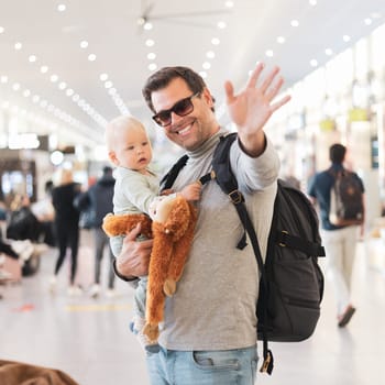 Father traveling with child, holding his infant baby boy at airport terminal waiting to board a plane waving goodby. Travel with kids concept