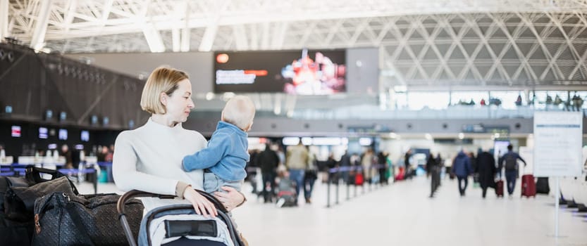 Motherat travelling with his infant baby boy child, walking, pushing baby stroller and luggage cart at airport terminal station. Travel with child concept