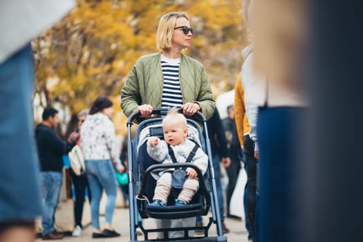 Mother waling and pushing his infant baby boy child in stroller in crowd of people wisiting sunday flea market in Malaga, Spain