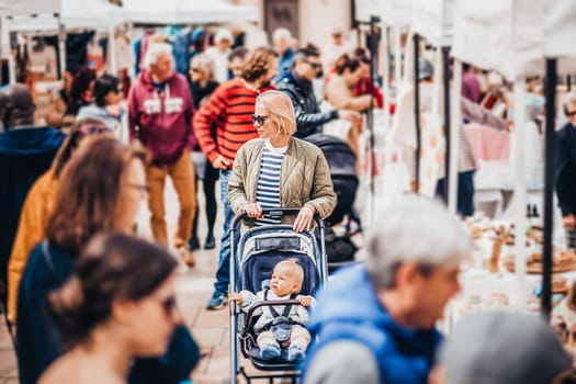 Mother waling and pushing his infant baby boy child in stroller in crowd of unrecognizable people wisiting sunday flea market in Malaga, Spain