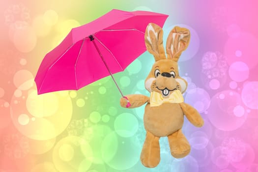 A cute brown plush bunny or rabbit with bow tie and pink umbrella over abstract rainbow background. Easter greeting card decoration element with space for design. Bright plush toy.