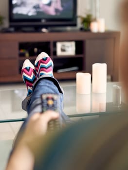 Young caucasian woman laying on sofa with colourful socks. She puts her feet on table and relaxes. The girl watches TV and holds remote control. Focus on socks