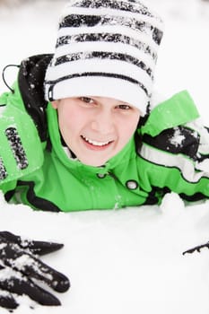 Friendly happy young boy with a charming smile playing in winter snow lying on his stomach looking up at the camera in a knitted beanie hat and warm green jacket