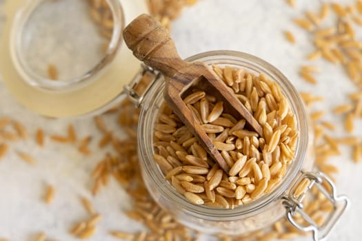Glass jar of raw dry rye grain with a wooden spoon on white table close up, top view. Healthy alternative low gluten seeds. Protein source for vegan and vegetarian diet.