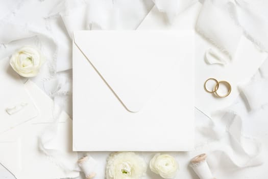 Square envelope near cream roses, white silk ribbons and wedding rings top view, wedding mockup. Romantic scene with pastel flowers flat lay. Valentines, Spring or Mothers day concept