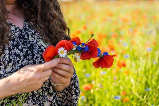 A long-haired curly woman weaves a wreath of wildflowers, poppies, daisies and cornflowers.